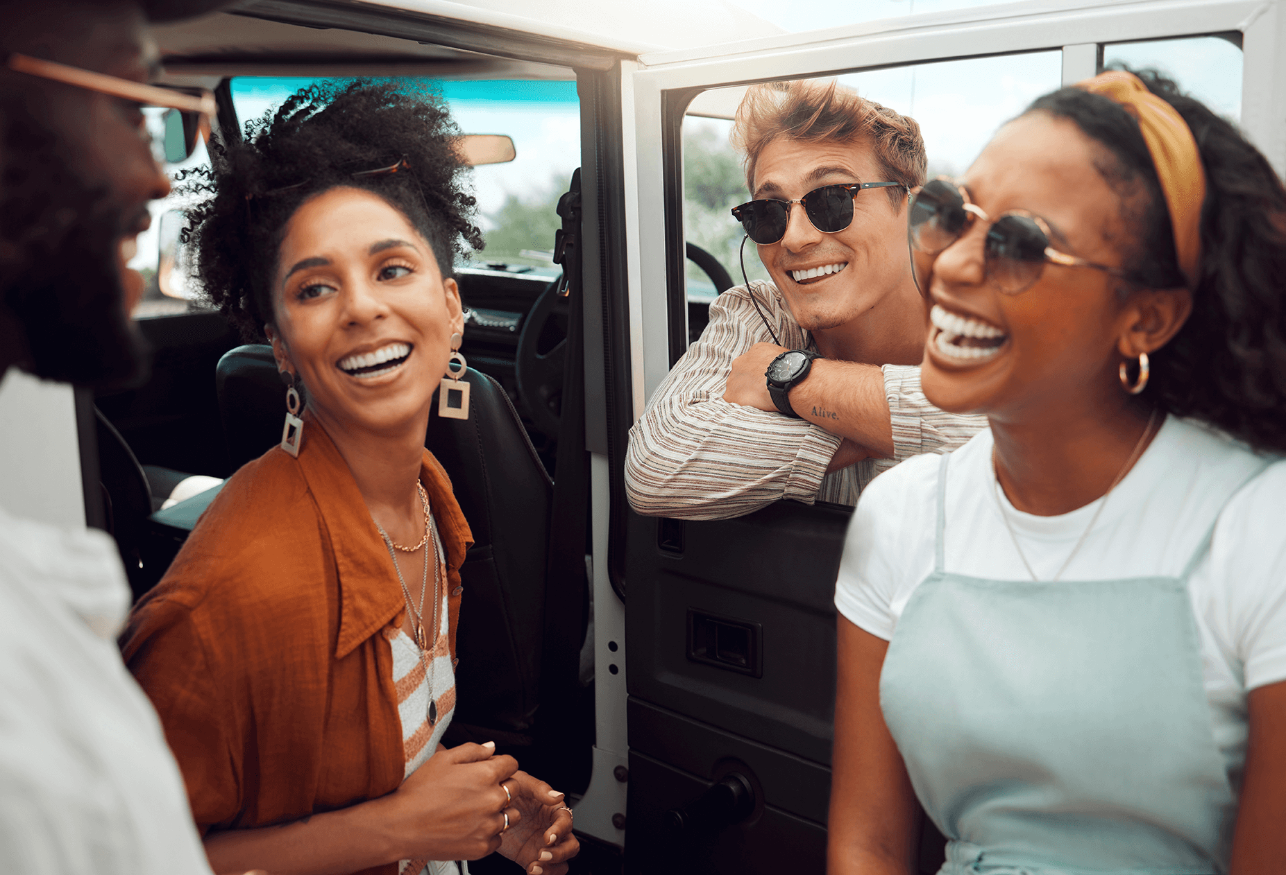 Group of friends laughing and talking by a car during a sunny road trip, representing friendship, travel adventures, and joyful outdoor experiences.