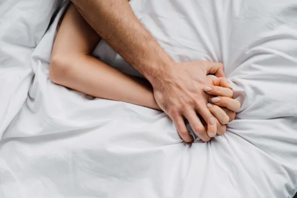 Intimate photo of a man's hand holding a woman's hand on white sheets.