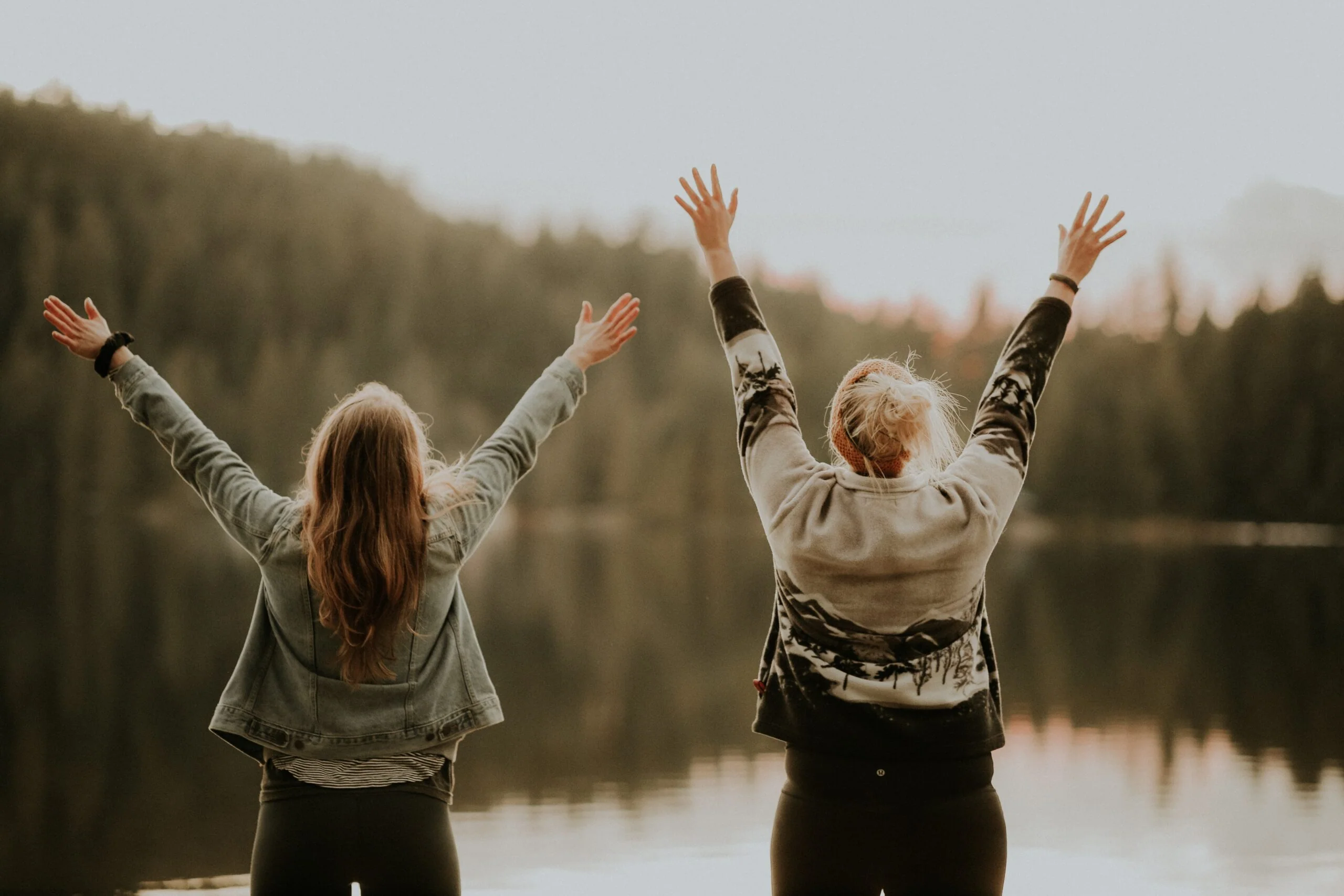 Two women outdoors with raised arms facing a lake and forest at sunset, symbolizing vitality, energy, and wellness benefits of NAD+ injections.
