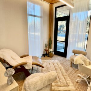 A therapy or relaxation room with a recliner, chairs with fluffy covers, a rug, plants, and large windows with sheer curtains.