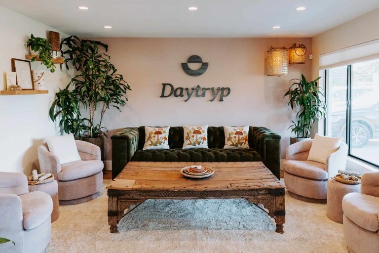 A lounge area with a green tufted sofa and pink swivel chairs arranged around a wooden coffee table. The Daytryp logo is on the wall behind the sofa, with large plants on either side.