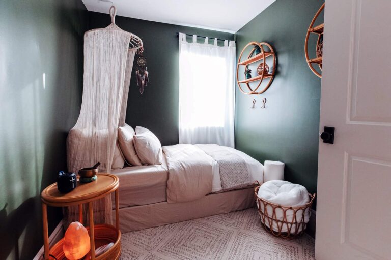 A small room with dark green walls, a bed with a canopy, a nightstand with a salt lamp, decorative shelves, a window with sheer curtains, and a basket.