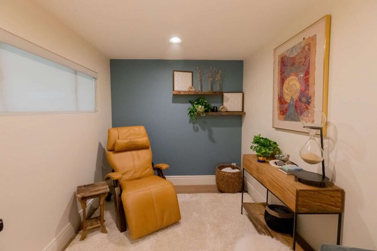 A room with a light blue accent wall. A tan leather reclining chair sits next to a wooden stool. A wooden console table on the right holds plants, books, and an hourglass, beneath a large framed abstract painting. Floating shelves with decor are on the blue wall.