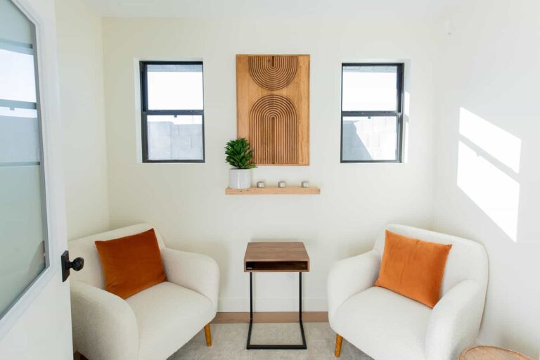 Two armchairs with orange pillows and a side table in a room with wooden wall art and windows, likely a therapy or waiting area.
