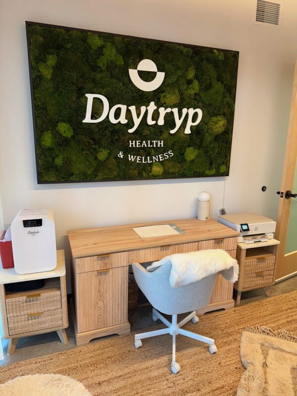 A room with a wooden desk, a chair with a fluffy cover, and a fuzzy chair. Above the desk is a large wall art piece with the Daytryp logo and "HEALTH & WELLNESS".
