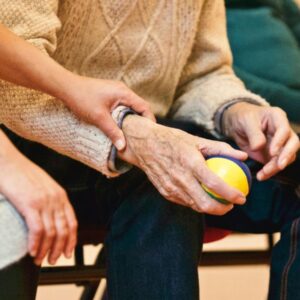 A caregiver gently holding the arm of an elderly person who is holding a colorful stress ball.