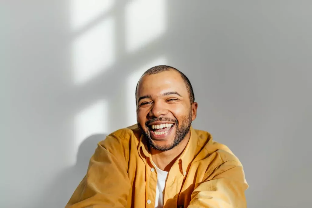 Bearded man in a yellow shirt laughing naturally, backlit by soft window light.