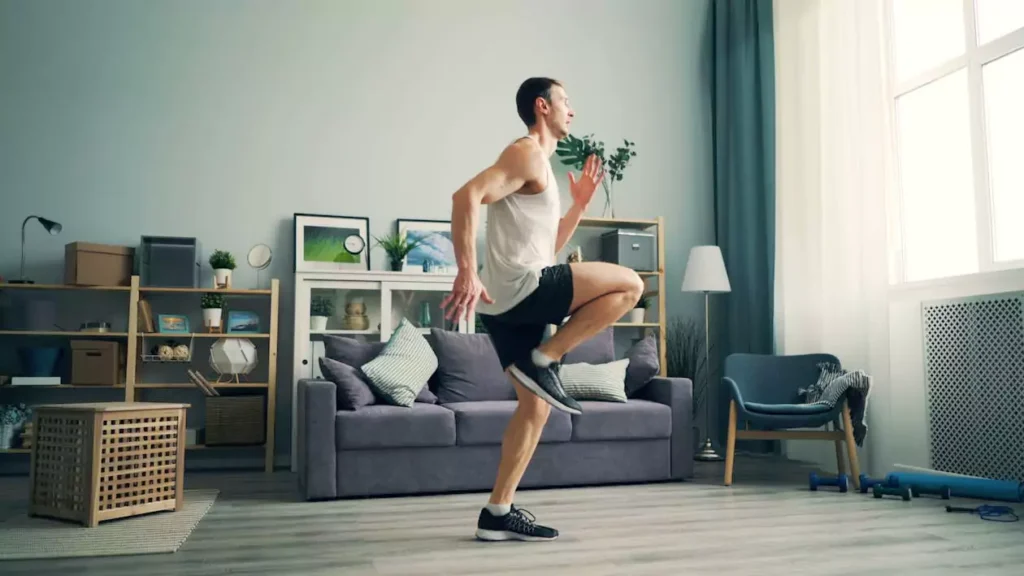 Man in a tank top and shorts doing a high-knee exercise at home in his living room.