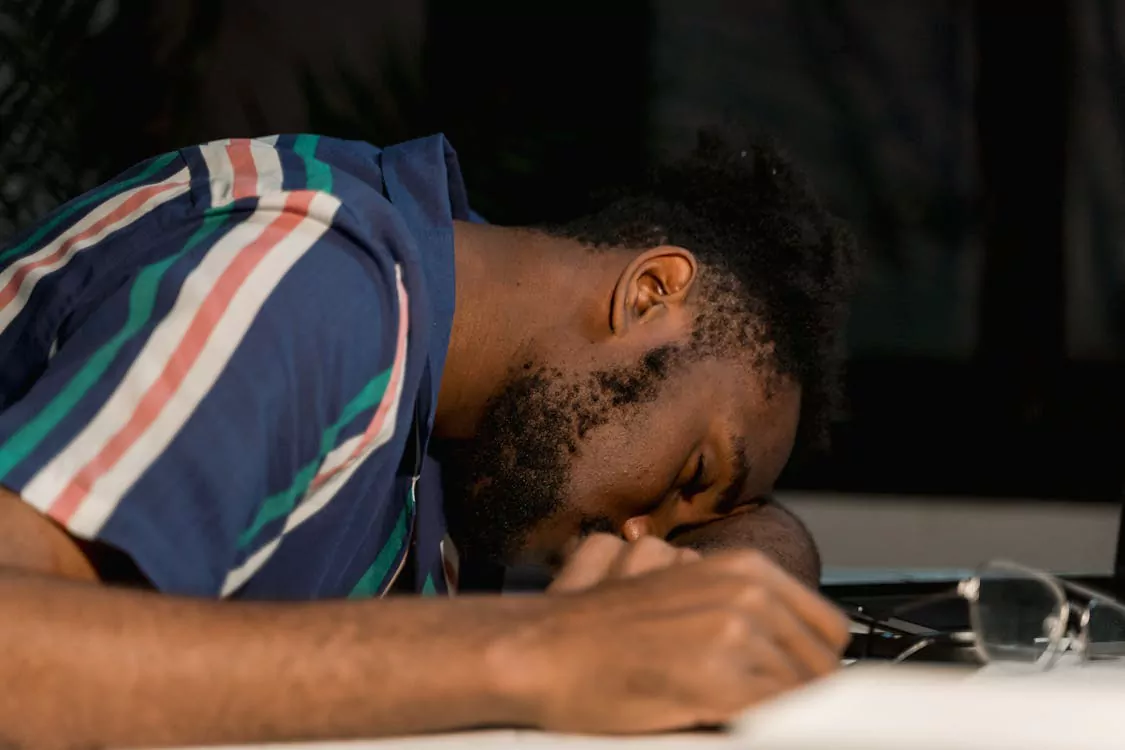 Young man in a striped shirt resting his head on his arm over a laptop, appearing exhausted.