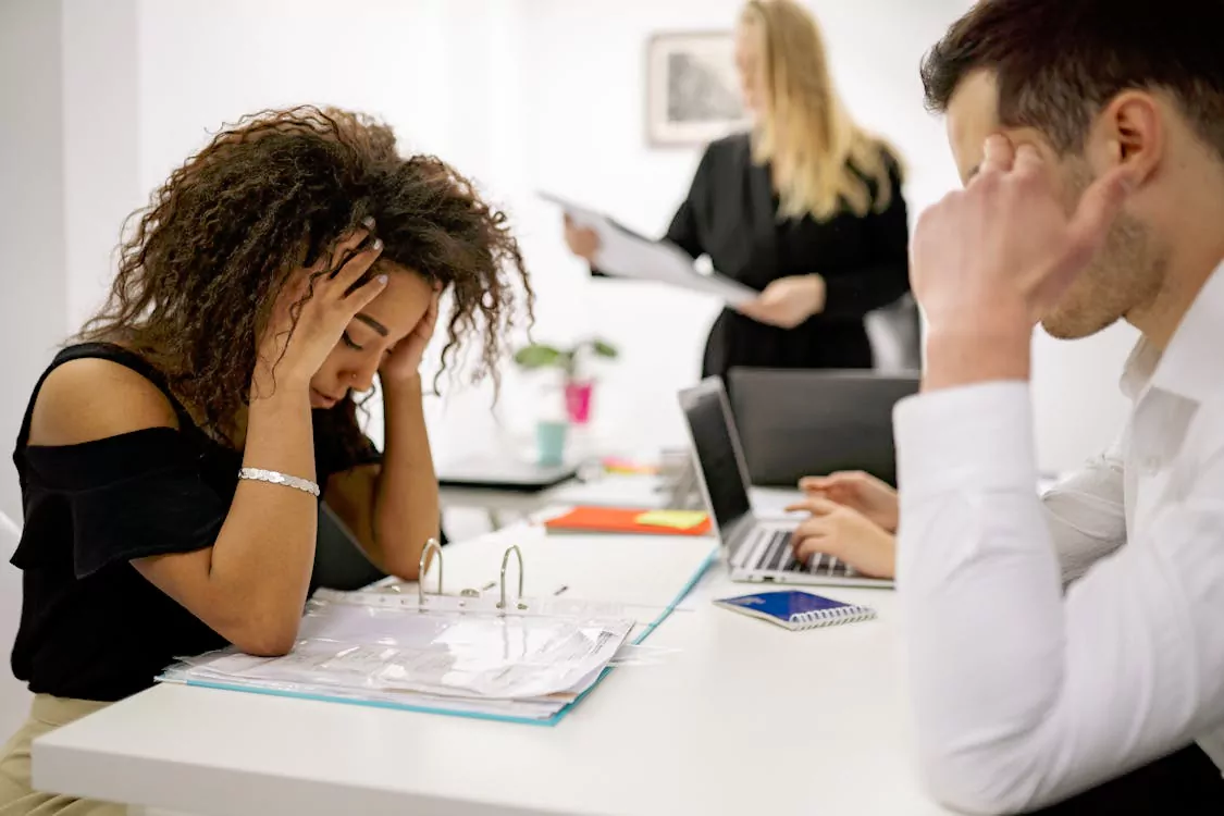 Stressed woman with curly hair holding her head at a cluttered office desk while colleagues work nearby.