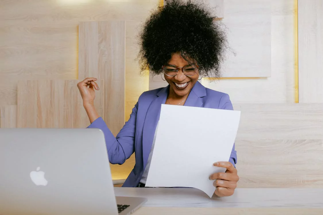 Woman in a purple blazer laughing joyfully while reading a document at her desk, laptop in front of her.