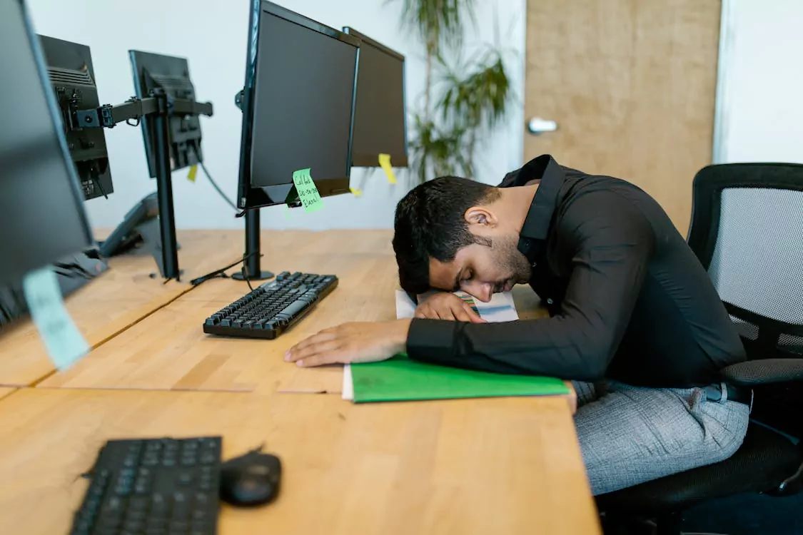 Man in a black shirt asleep at his desk with his head resting on his arms, in front of dual monitors.