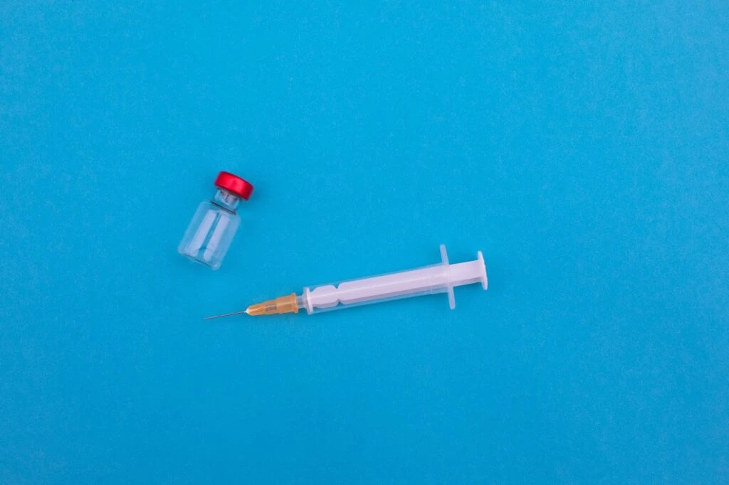 A glass vial with a red cap and a syringe on a blue surface.