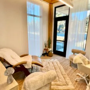 A therapy or relaxation room with a recliner, chairs with fluffy covers, a rug, plants, and large windows with sheer curtains.