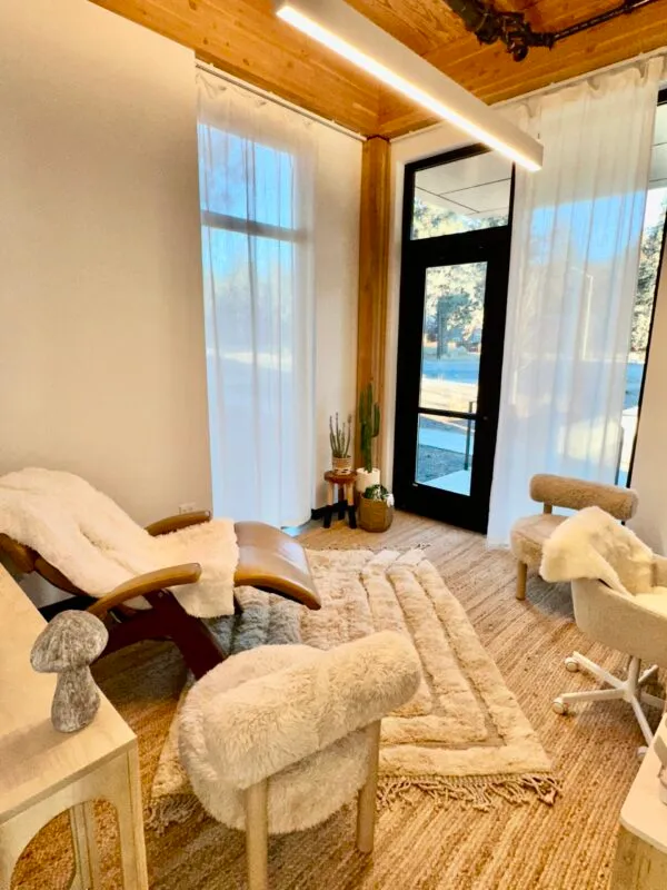 A therapy or relaxation room with a recliner, chairs with fluffy covers, a rug, plants, and large windows with sheer curtains.