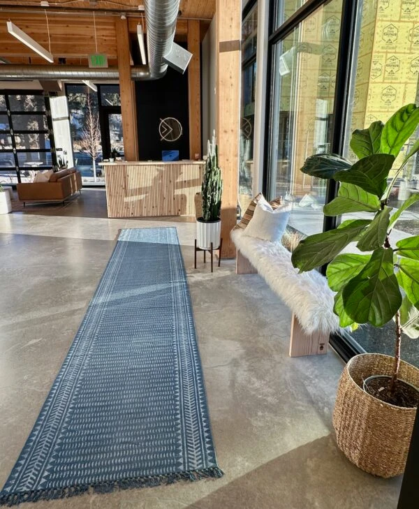 A modern entrance or lobby area with a long patterned rug, a wooden reception desk, a fluffy bench, and large windows.
