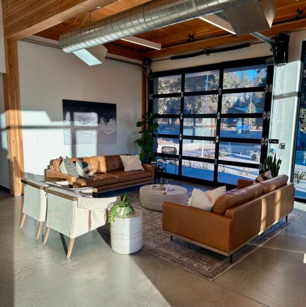A modern lounge area with concrete floors, exposed ducts, leather couches, armchairs, and large windows resembling garage doors.
