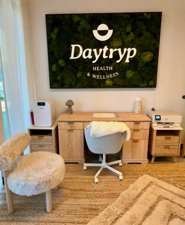 A room with a wooden desk, a chair with a fluffy cover, and a fuzzy chair. Above the desk is a large wall art piece with the Daytryp logo and "HEALTH & WELLNESS".