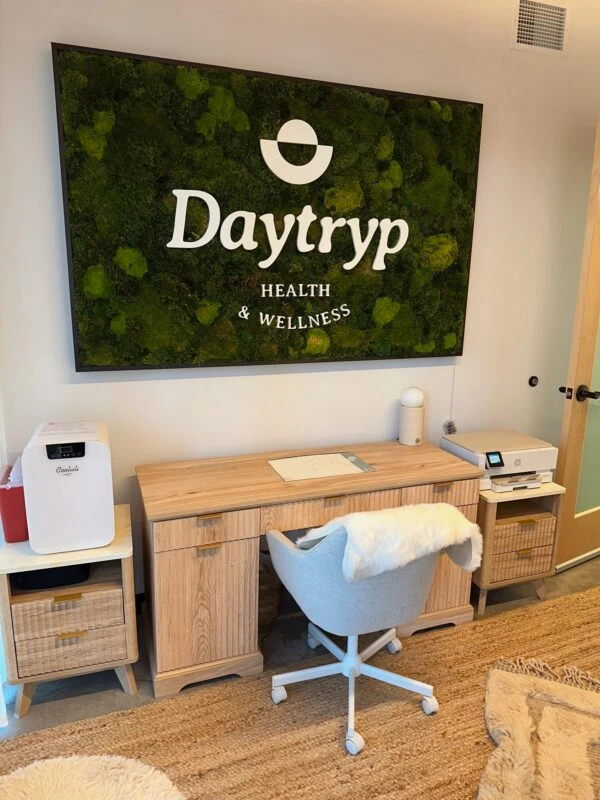 A room with a wooden desk, a chair with a fluffy cover, and a fuzzy chair. Above the desk is a large wall art piece with the Daytryp logo and "HEALTH & WELLNESS".