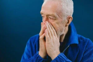 An older man covers his mouth and nose with his hands, looking distressed.